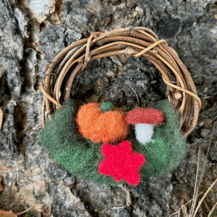 Miniature wreath sits at the base of a birch tree. On its bottom is green felting wool topped with an orange pumpkin, a mushroom, and a red maple leaf in center.