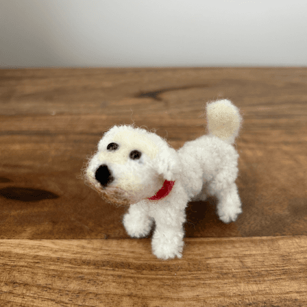 A white miniature schnauzer posed standing made out of thick chenille pipe cleaner and wool with glass bead eyes. It has a dirty brown mouth and a red collar. It is facing towards the bottom left of the image.