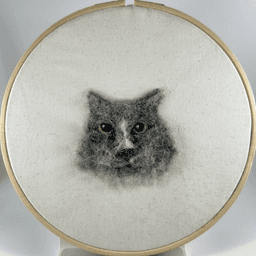 Wool portrait of grey cat Max in an 8 inch embroidery frame. Max has green eyes and white marks on his nose.