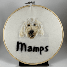 Needle felted portrait of a white goldendoodle with its mouth open in a relaxed smile and her name, Mamps written below.