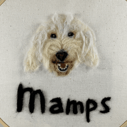 Close up of a needle felted portrait of a white goldendoodle with its mouth open in a relaxed smile and her name, Mamps written below.