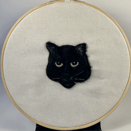 Needle felted portrait of a black cat with green eyes and a flat expression.