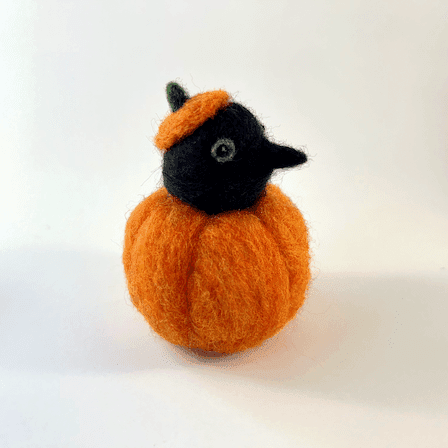 A needle felted black corvid borb pokes its head out of an orange pumpkin wearing a pumpkin stem hat. The borb faces right. Its eyes are lined in grey with a reflection.