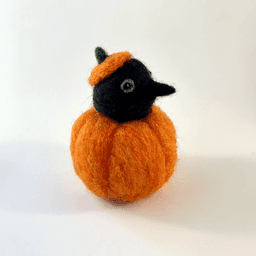 A needle felted black corvid borb pokes its head out of an orange pumpkin wearing a pumpkin stem hat. The borb faces right. Its eyes are lined in grey with a reflection.