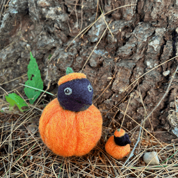 Two needle felted borbs sit at the base of a pine tree on some pine needles. They’re both black corvids with their heads sticking out of a pumpkin with a pumpkin stem hat. The left borb is a couple inches tall and wide, but the right is a miniature version that could fit on a US penny.