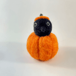 A needle felted black corvid borb pokes its head out of an orange pumpkin wearing a pumpkin stem hat. The borb faces directly towards the camera. Its eyes are lined in grey with a reflection.