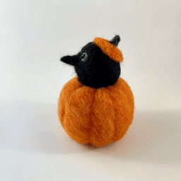 A needle felted black corvid borb pokes its head out of an orange pumpkin wearing a pumpkin stem hat. The borb faces directly left. Its eyes are lined in grey with a reflection.