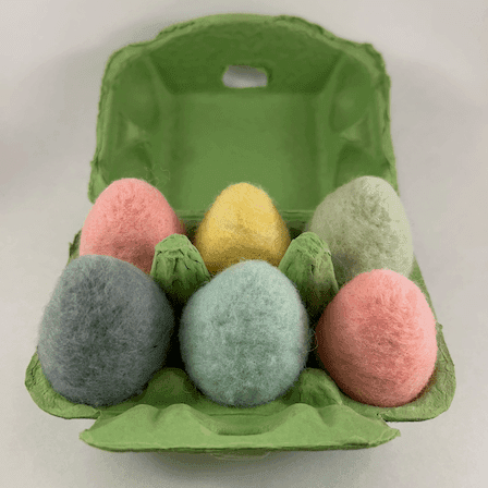 A half dozen pastel colored needle-felted eggs sit in a green egg carton. The eggs are shades of pink, yellow, and blue.