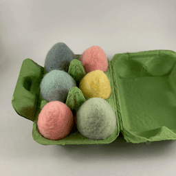 A half dozen pastel colored needle-felted eggs sit in a green egg carton. The eggs are shades of pink, yellow, and blue.