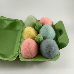 A half dozen pastel colored needle-felted eggs sit in a green egg carton. The eggs are shades of pink, yellow, and blue.