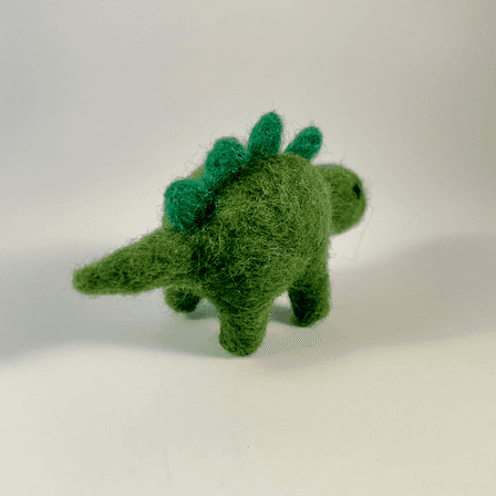 A needle-felted green stegasaurus faces away from the camera 1/4 to the right.