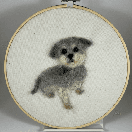 A wool portrait of Sylvie the grey and white yorkie poodle mix sitting 3/4 towards the right, her face looking slightly left towards the camera, her eyes expressive, round white beard and paws slightly saliva-stained, ears and tail dark grey, framed in an 8 inch embroidery hoop.