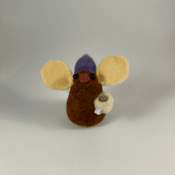 A needle-felted chocolate colored mouse wearing a purple hat with blue pom pom and holding a cream colored mug facing towards the camera.