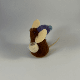 A needle-felted chocolate colored mouse wearing a purple hat with blue pom pom and holding a cream colored mug facing towards the left.