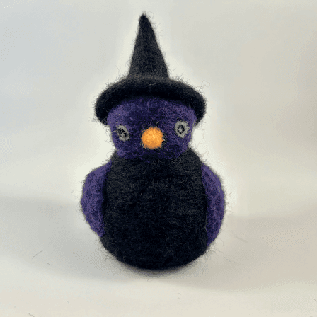 A needle-felted witch borb faces forward. It has a purple face, purple wings, light green eyes and an orange beak. The witch has a black body and wears a black witch cap.