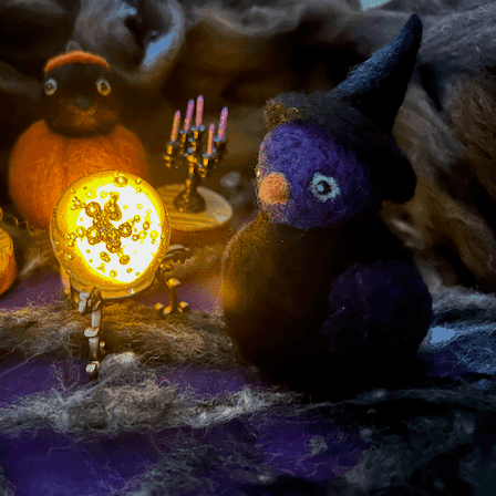 A needle-felted witch borb faces 3/4 to the left. It has a purple face, purple wings, light green eyes and an orange beak. The witch has a black body and wears a black witch cap. It sits in a spooky scene with a glowing crystal ball to the left, a purple candelabra behind, and a Pumpkin Borb in the background. The floor is purple and cobwebs and the background is dingy.