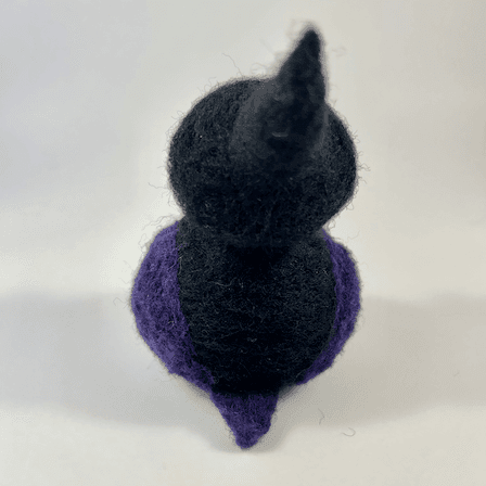 A needle-felted witch borb faces away from the camera. It has a purple tail and purple wings. The witch has a black body and wears a black witch cap.