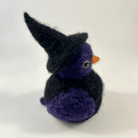 A needle-felted witch borb faces right. It has a purple face, purple wings, light green eyes and an orange beak. The witch has a black body and wears a black witch cap.