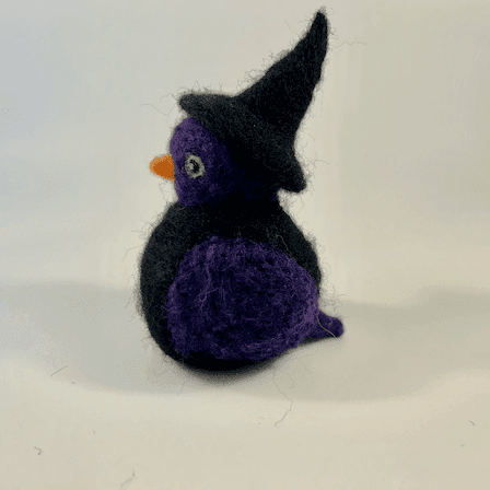 A needle-felted witch borb faces left. It has a purple face, purple wings, light green eyes and an orange beak. The witch has a black body and wears a black witch cap.