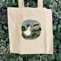Close up of canvas tote bag with a wizard goose felted on - wearing a purple hat and shoes and honking standing among shrubs and flowers with a tiny ladybug friend.