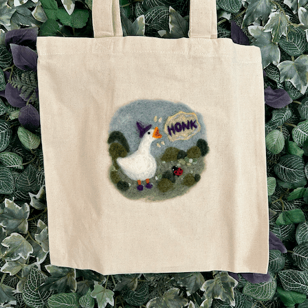Close up of canvas tote bag with a wizard goose felted on - wearing a purple hat and shoes and honking standing among shrubs and flowers with a tiny ladybug friend.