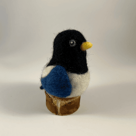 A needle-felted yellow-billed magpie borb sits on a circle of cut wood, facing 3/4 towards the right.