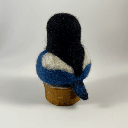 A needle-felted yellow-billed magpie borb sits on a circle of cut wood, facing away from the camera 1/4 towards the left.