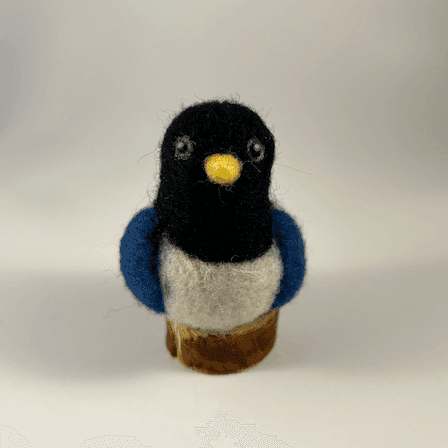 A needle-felted yellow-billed magpie borb sits on a circle of cut wood, facing directly towards the camera.