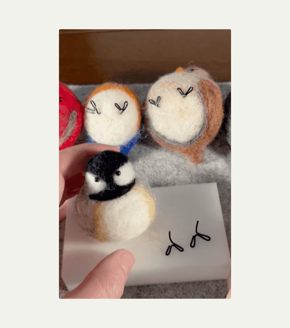 In the background, several needle felted borbs sit upturned revealing their bases and black wire feets. In front, a needle felted black-capped chickadee sits on a foam felting pad next to two wire borb feets.