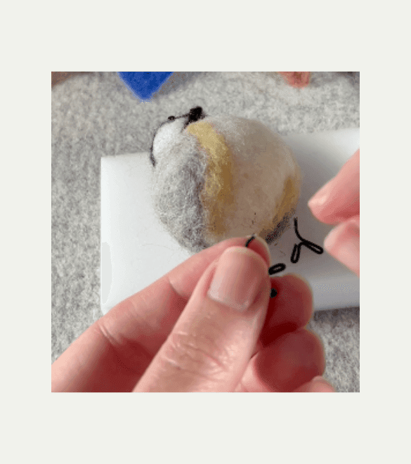 With an upturned black-capped chickadee lying on a foam felting pad in the background, a partially obscured left hand holds a borb feets with the so-called leg tip bent slightly.