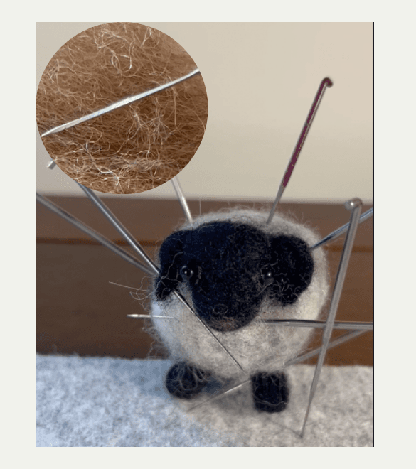 A needle-felted grey horned sheep with a black face has seven felting needles stuck inside it from several different directions. A circular inlay in the upper left shows a close up of a felting needle with barbs visible.