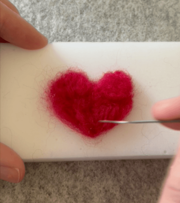 Close up of a heart shape being needle felted into a foam block. A mostly obscured hand is holding onto the block on the left.