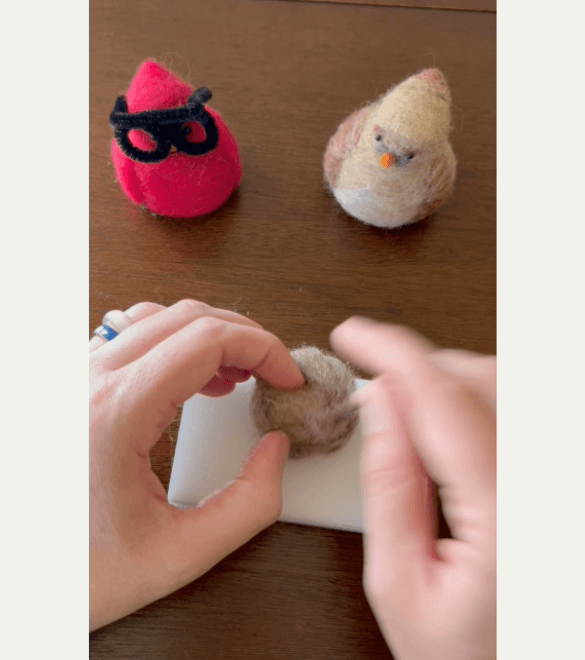 While a pair of needle felted Northern cardinals watch, a left hand with fingers curled holds a needle felted ball while a right hand pokes the ball with a felting needle.