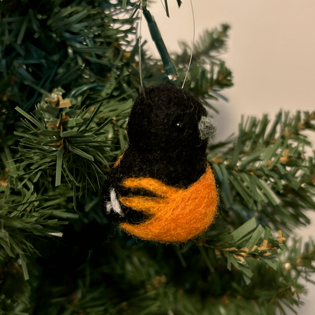 A close up of a needle-felted Baltimore oriole ornament, hanging on a tree, facing 1/4 to the right.