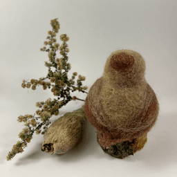 A female needle-felted Northern cardinal sits on a half-inch circle of cut branch, facing away from the camera 1/4 towards the right. There is a spray of goldenrod and a milkweed pod husk in the background. Her body is buff cream colored with wine-stained wings and tail, and wine-stained crest.