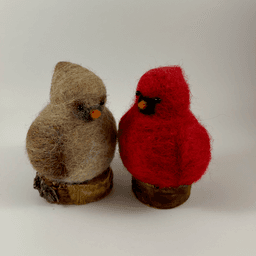 A pair of male and female Northern cardinals, scultped in needle felt, sit on half inch circle sections of branches, facing 1/4 inch towards each other. The female is a buff cream color with wine-stained wings, wine-stained eyebrows, a grey goatee and orange beak. The male is a cardinal red color with a black goatee and orange beak. Their crests are short and round.