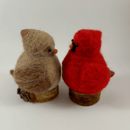 A pair of male and female Northern cardinals, scultped in needle felt, sit on half inch circle sections of branches, facing directly towards each other in profile. The female is a buff cream color with wine-stained wings, wine-stained eyebrows, a grey goatee and orange beak. The male is a cardinal red color with a black goatee and orange beak. Their crests and tail points are short and round.