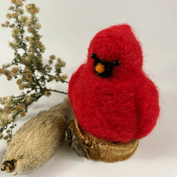 A male needle-felted Northern cardinal sits on a half-inch circle of cut branch, facing 1/4 towards the left. There is a spray of goldenrod and a milkweed pod husk in the background. His body is a bright red color, and his goatee is black, his beak orange. His crest is short and round.