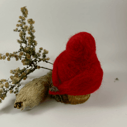 A male needle-felted Northern cardinal sits on a half-inch circle of cut branch, facing away from the camera 1/4 towards the right. There is a spray of goldenrod and a milkweed pod husk in the background. His body is a bright red color, the tip of his tail grey. His crest is short and round.