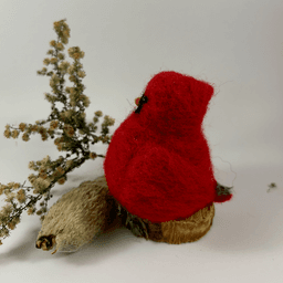 A male needle-felted Northern cardinal sits on a half-inch circle of cut branch, facing towards the left in profile. There is a spray of goldenrod and a milkweed pod husk in the background. His body is a bright red color, and his goatee is black, his beak orange. His crest is short and round.