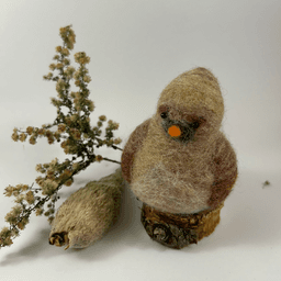 A female needle-felted Northern cardinal sits on a half-inch circle of cut branch, facing 1/4 towards the left. There is a spray of goldenrod and a milkweed pod husk in the background. Her body is a buff cream color, her wings and eyebrows are wine-stained, her goatee is grey, her beak orange. His crest is short and round.