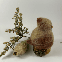 A female needle-felted Northern cardinal sits on a half-inch circle of cut branch, facing towards the right in profile. There is a spray of goldenrod and a milkweed pod husk in the background. Her body is a buff cream color, her wings, crest and tail are wine-stained, her beak orange. His crest is short and round.