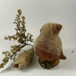 A female needle-felted Northern cardinal sits on a half-inch circle of cut branch, facing away from the camera 1/4 towards the left. There is a spray of goldenrod and a milkweed pod husk in the background. Her body is buff cream colored with wine-stained wings and tail, and wine-stained crest.